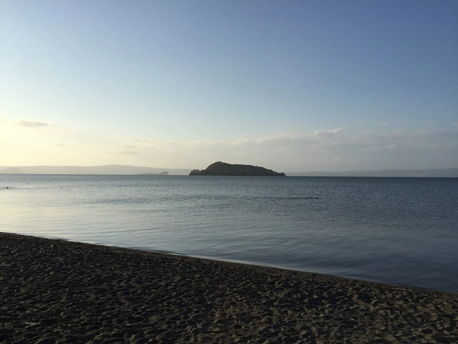 Spiaggia di sabbia scura vulcanica sul lago di Bolsena con l'acqua calma e un'isola verde all'orizzonte nella luce del tardo pomeriggio